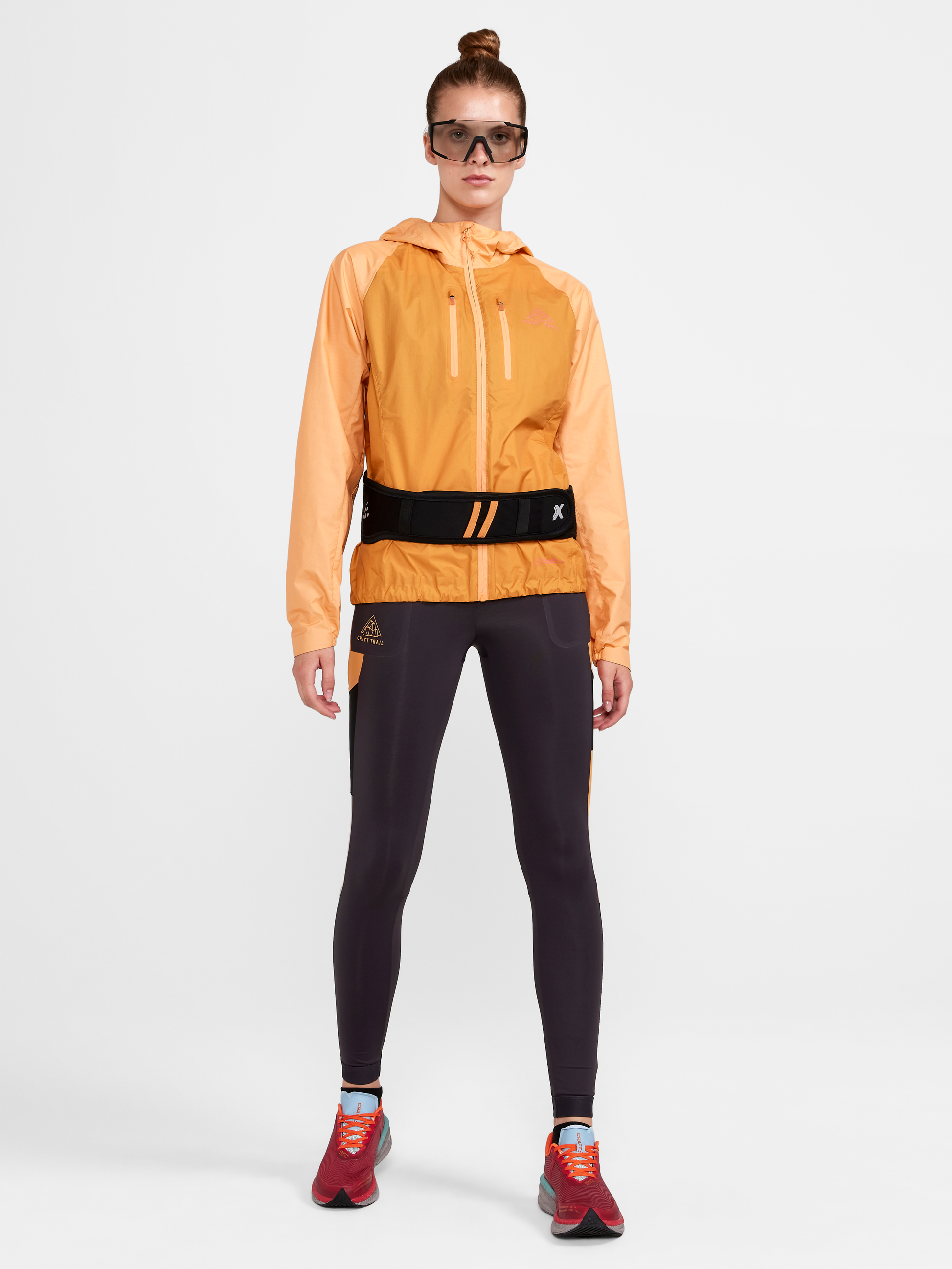 Woman models a light orange hooded running jacket, black running tights, and a black running waist belt.
