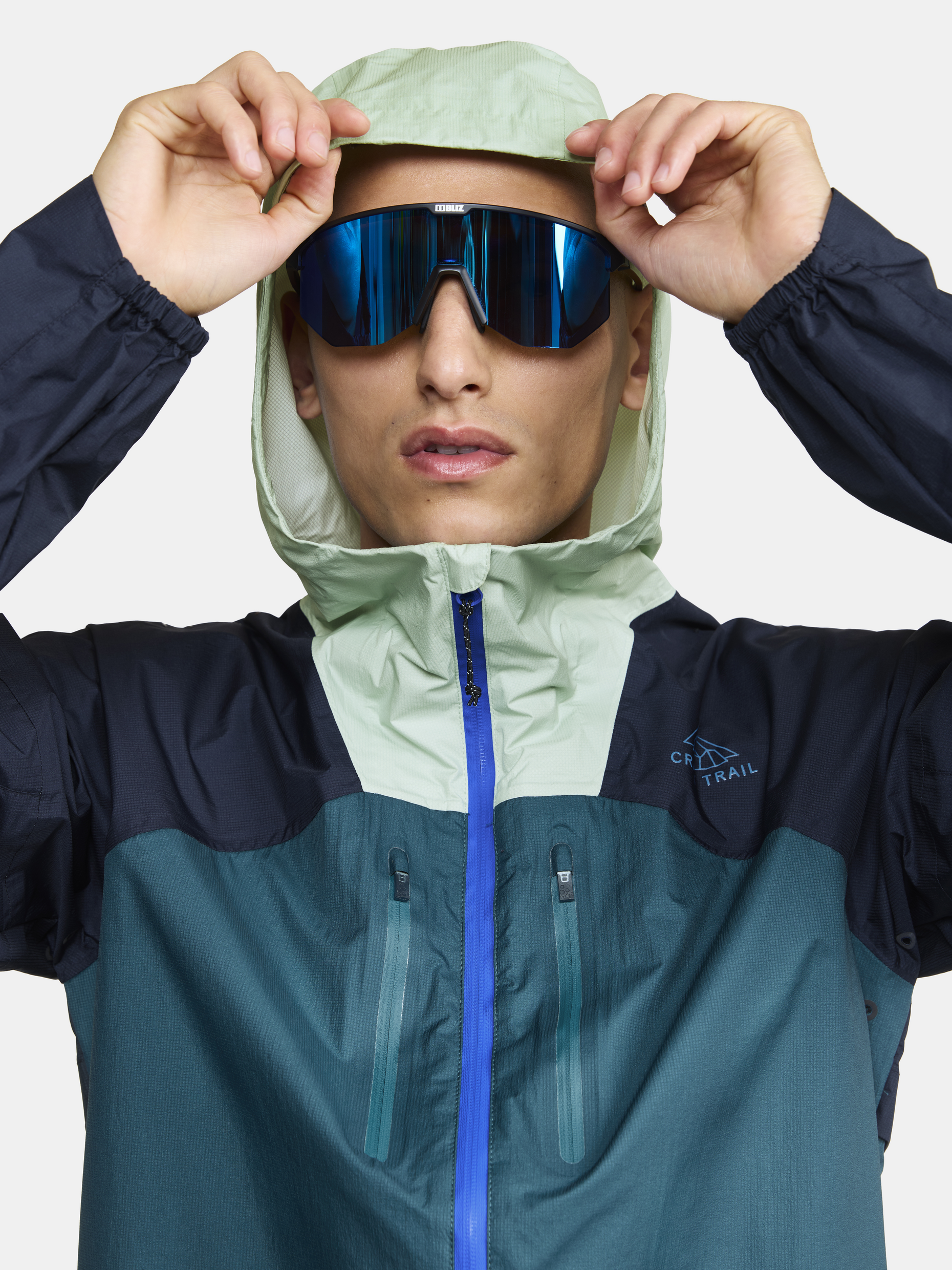 Man adjusts the hood of his teal and navy blue CRAFT Sportswear running jacket.