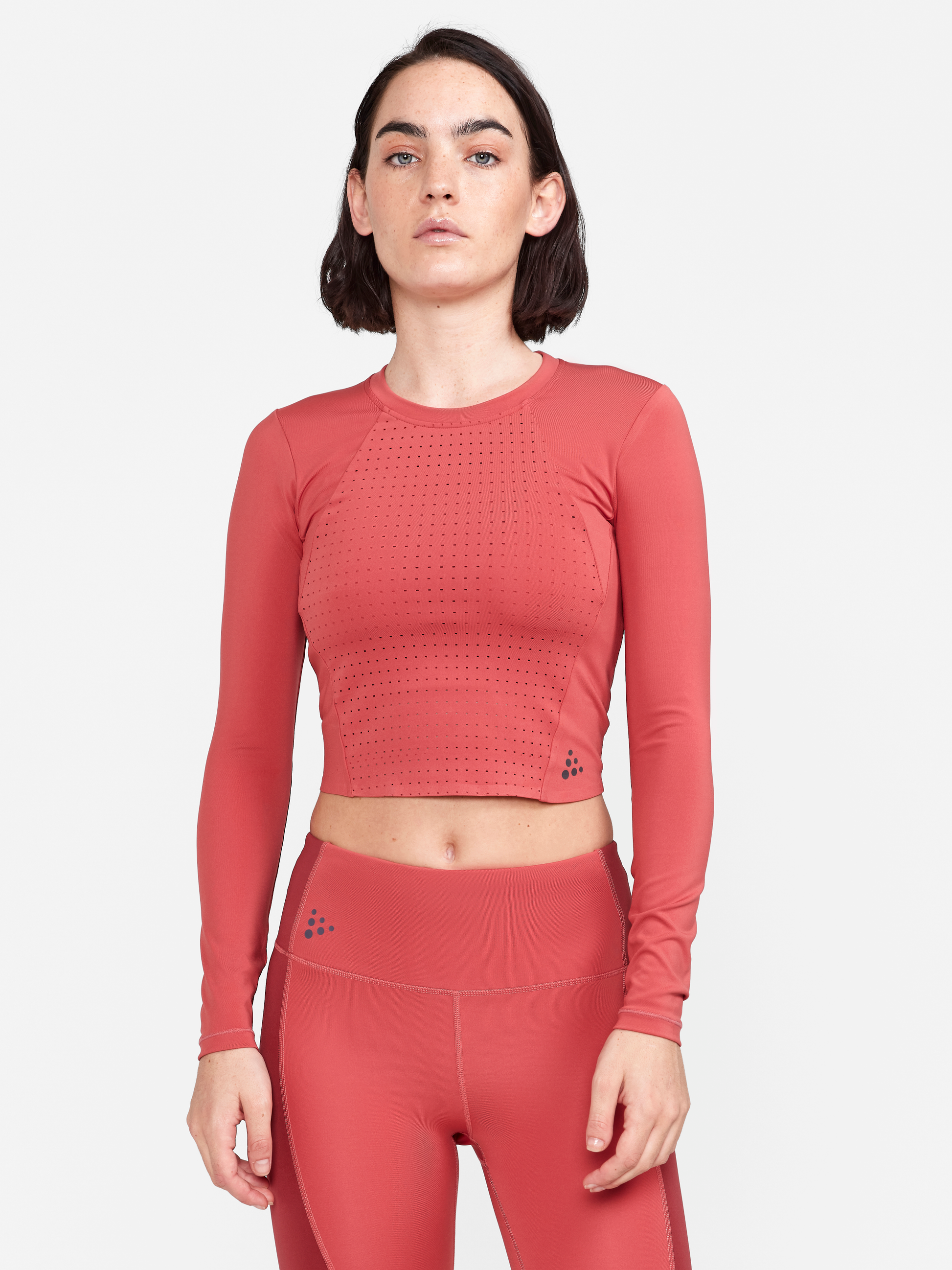 Woman models a coral pink long-sleeve athletic crop top with laser-cut ventilation details and matching leggings.