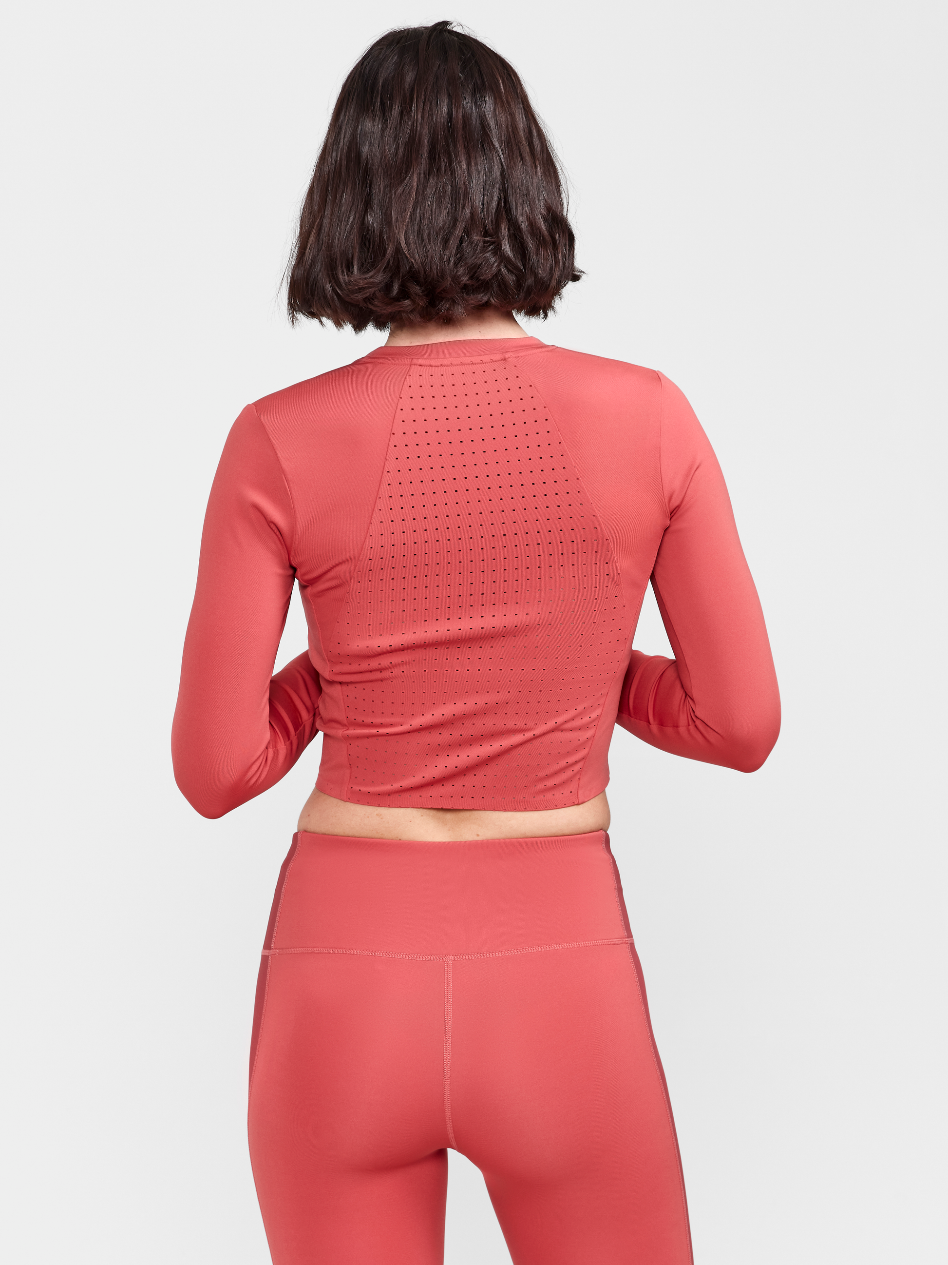 Woman models a coral pink athletic long-sleeve crop top with breathable mesh back panel.