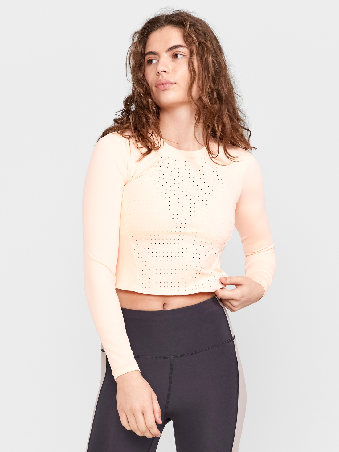 Woman models a peach cropped athletic top with laser-cut perforations, showcasing its breathable design.