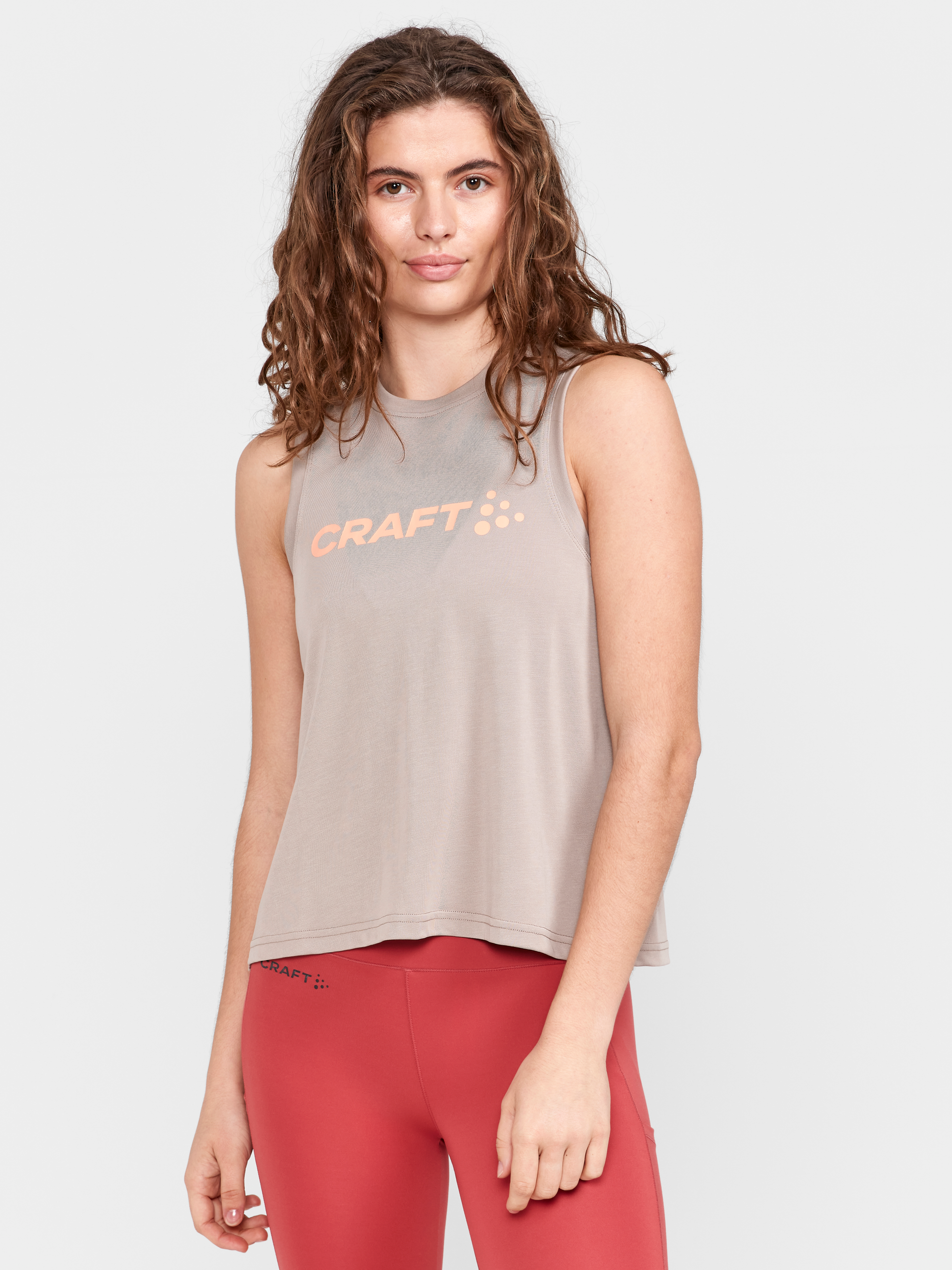 Woman wears a beige sleeveless Craft sportswear top and red training leggings.