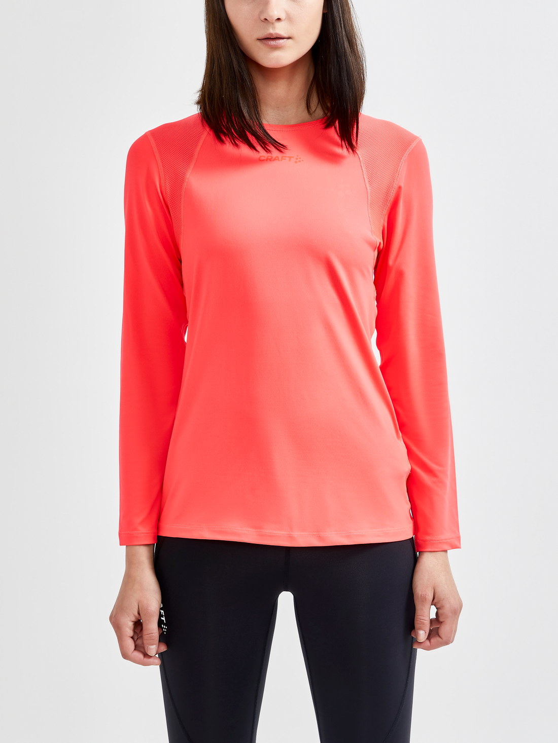 Woman models a bright coral long-sleeve athletic shirt with breathable mesh panels on the shoulders and sides. 
