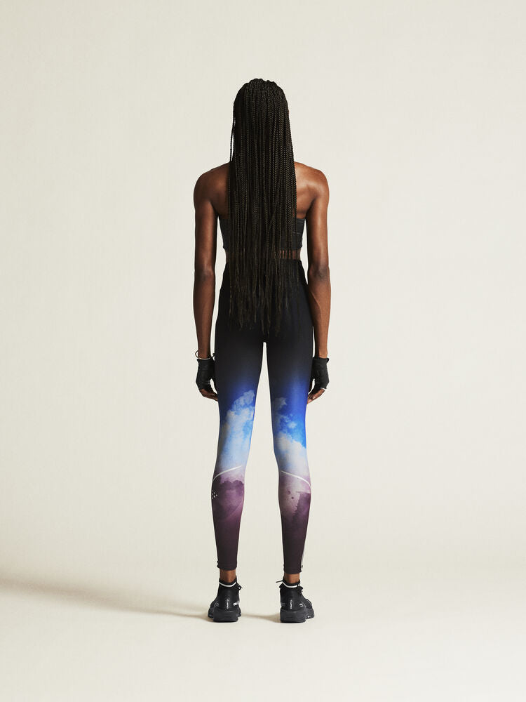 Woman models black Craft high-waisted running leggings with blue and purple ombre cloud design. 
