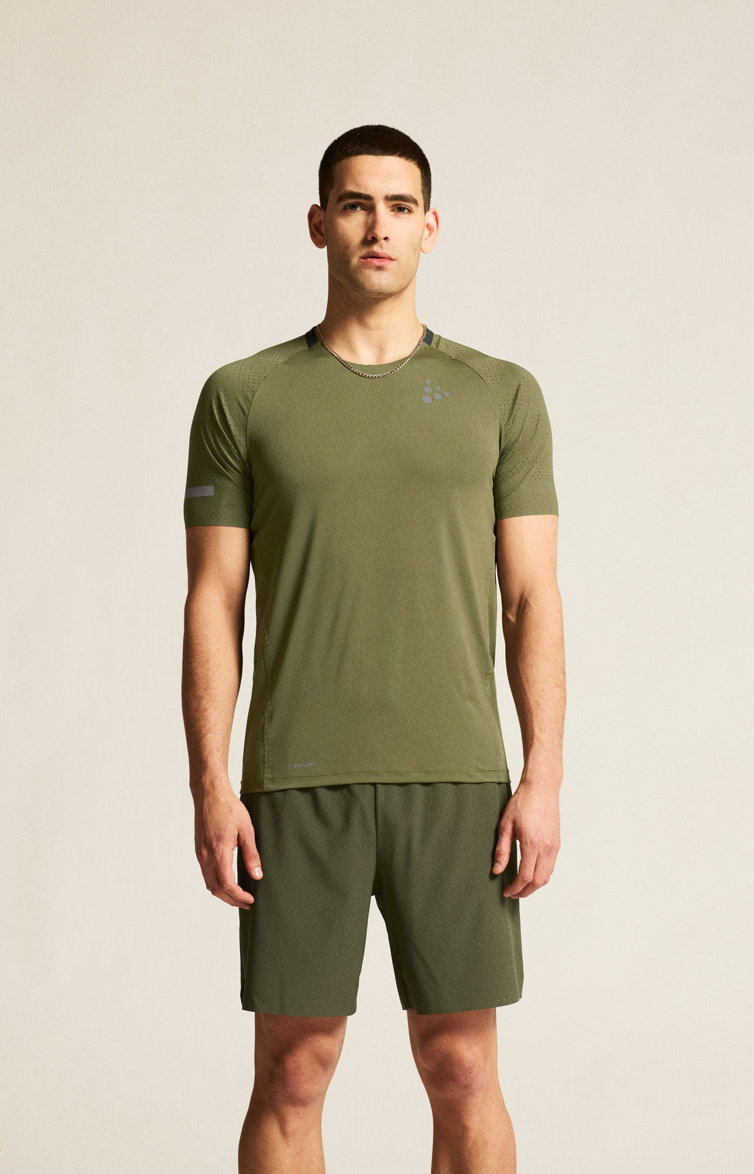 Man models an olive green athletic t-shirt and matching shorts against a beige background. 
