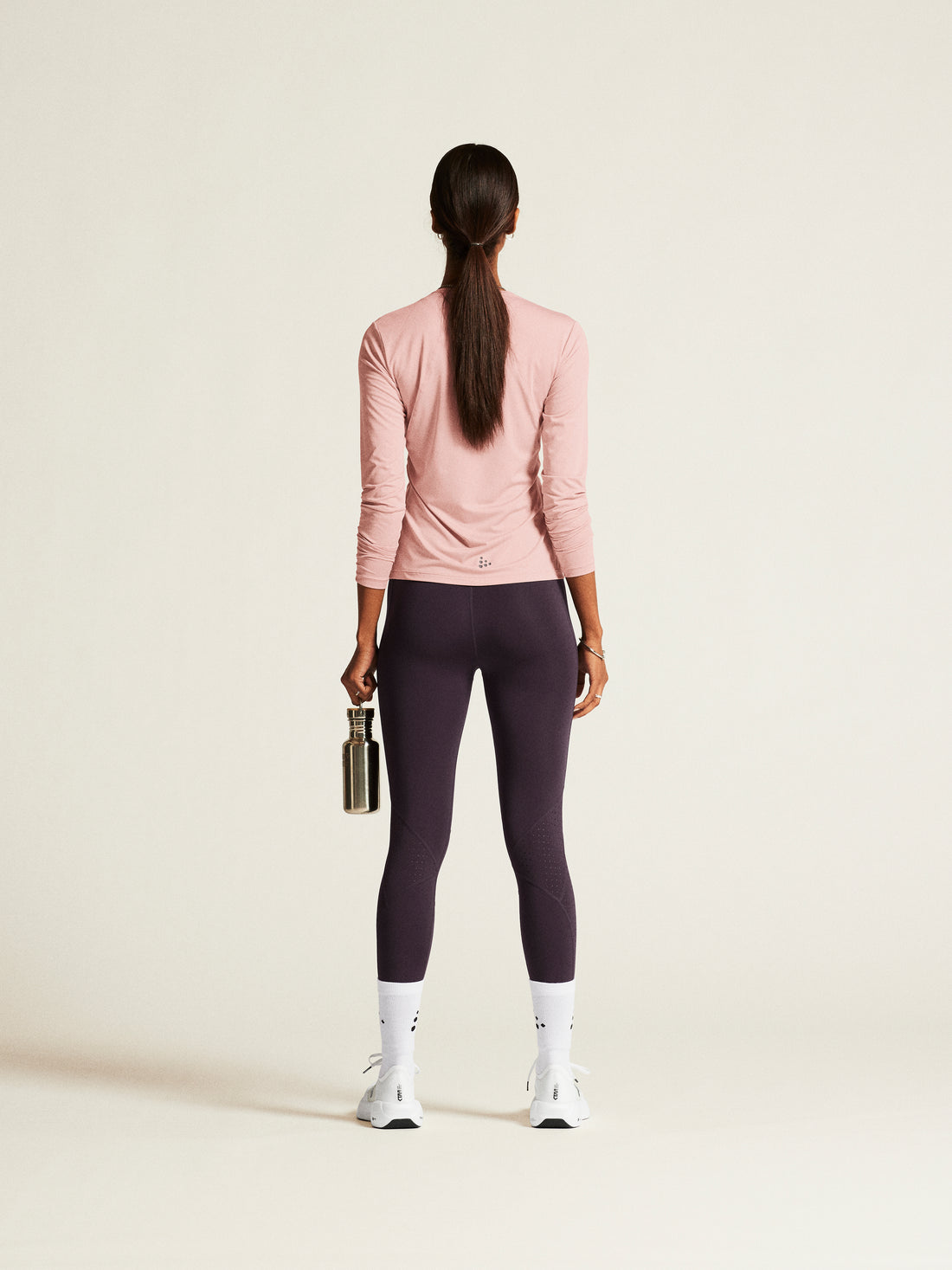 Woman models a pink athletic long-sleeve shirt, purple running tights, and white sunglasses while holding a reusable water bottle. 
