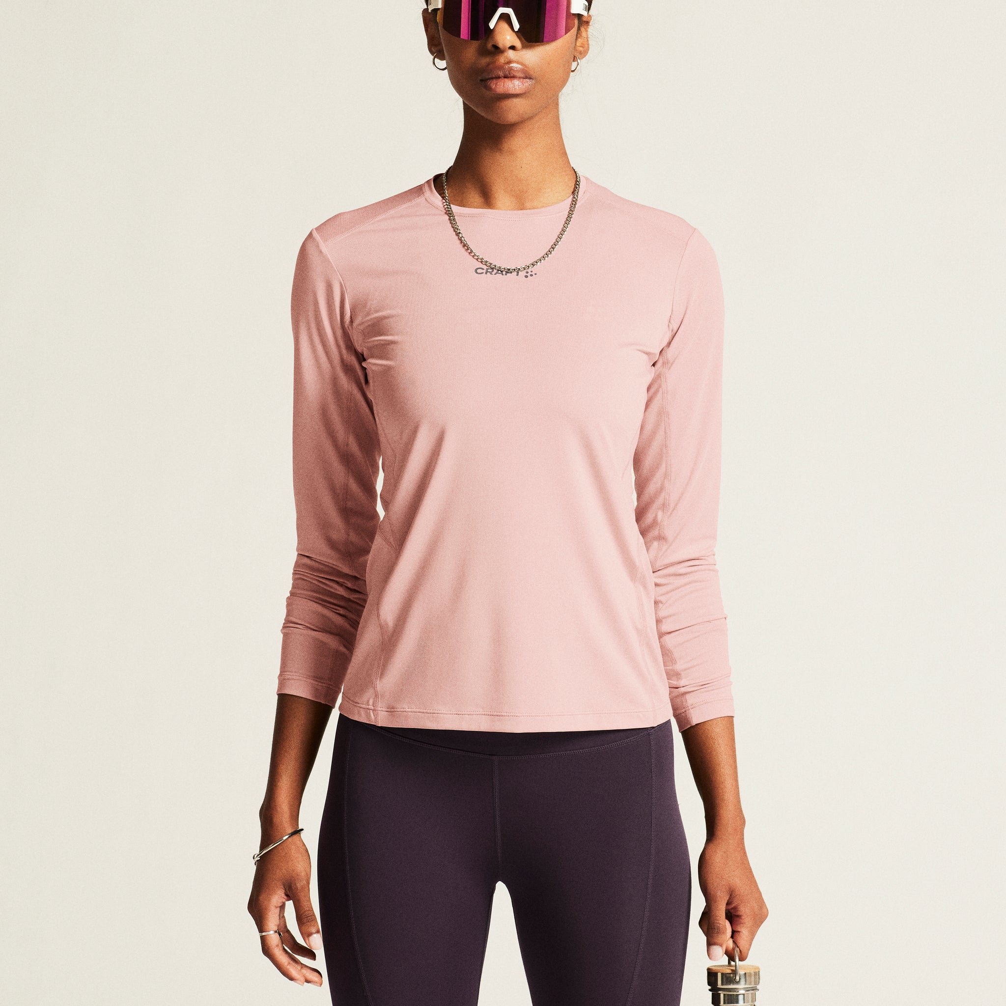 Woman models a pink athletic long-sleeve shirt, purple running tights, and white sunglasses while holding a reusable water bottle. 

