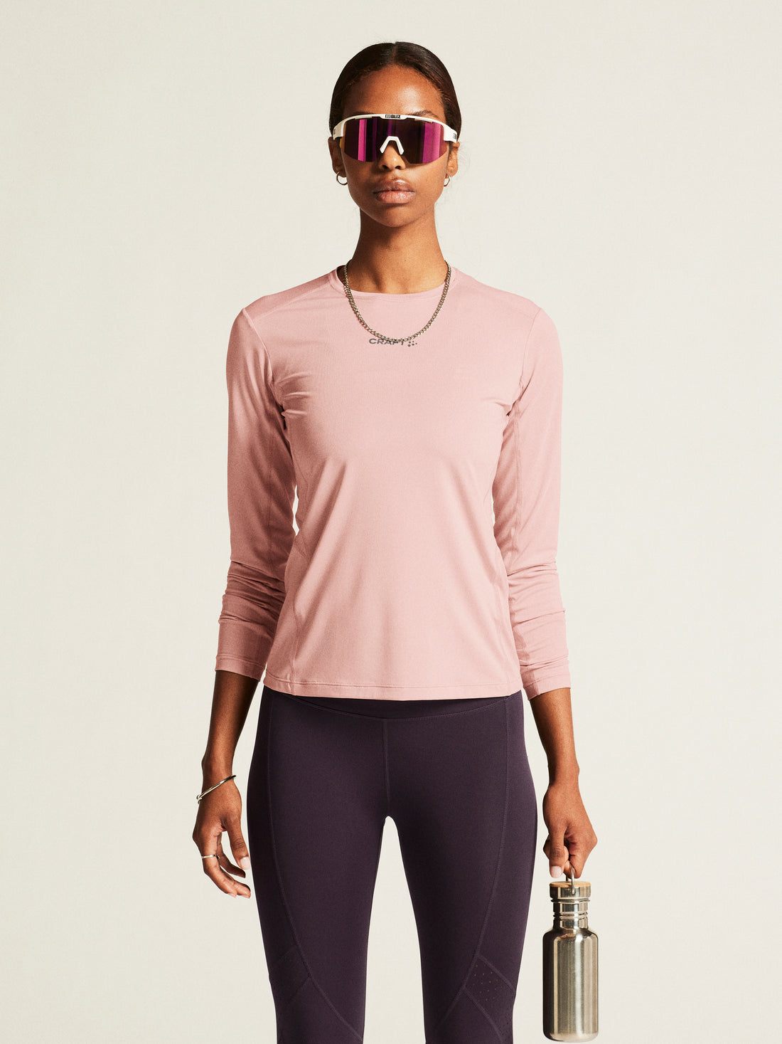 Woman models a pink athletic long-sleeve shirt, purple running tights, and white sunglasses while holding a reusable water bottle. 
