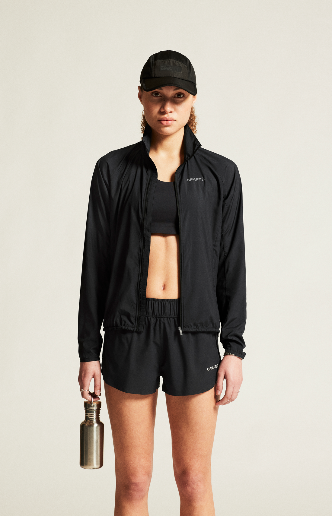 Woman holds reusable water bottle, wearing black Craft Sportswear running jacket, shorts, and cap. 
