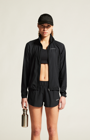 Woman holds reusable water bottle, wearing black Craft Sportswear running jacket, shorts, and cap. 

